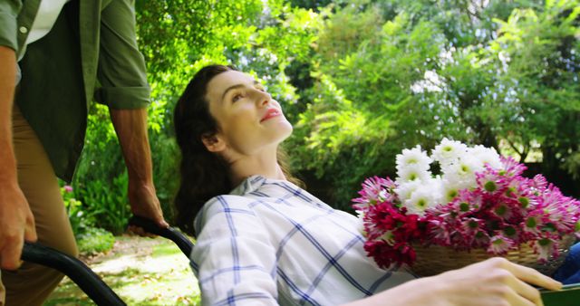 This vibrant image of a woman relaxing in a wheelbarrow while holding a basket of flowers is perfect for advertisements promoting gardening activities and outdoor relaxation. It could also be used in lifestyle magazines and well-being blogs to illustrate content about connecting with nature, taking leisurely outdoor breaks, or showcasing a serene and joyful moment in a garden setting.