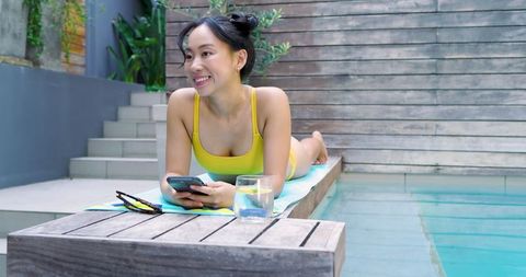 Asian woman relaxing by pool with smartphone in yellow swimwear