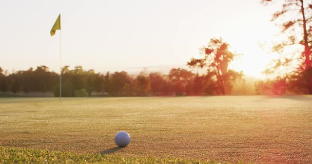 A close-up shot of a golf ball positioned near a hole with the flag still in place on a golf course. The scene is illuminated by warm, early morning sunlight, casting long shadows across the grass. This image is ideal for promoting golf courses, illustrating golf-related articles, or emphasizing the tranquility and beauty of early morning sports activities.