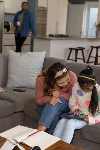 Mother and daughter using tablet on sofa while father watching from open-plan kitchen
