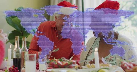Senior couple celebrating holiday dinner wearing santa hats with purple world map overlay