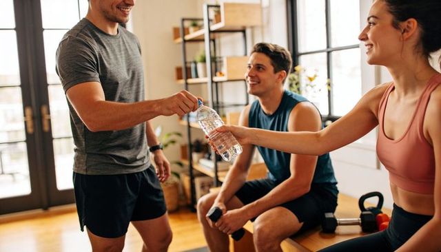 Friends exchanging water bottle in sunlit home gym after strength training session