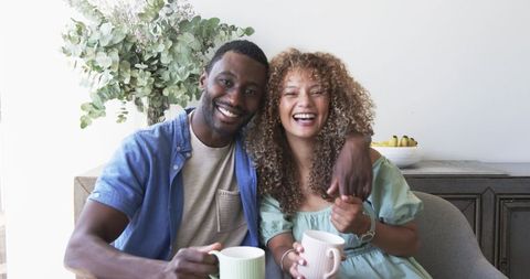 Smiling couple on sofa holding pastel mugs enjoying cozy morning together at home