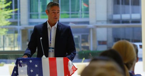 Professional speaker addressing an audience at a business seminar, with American flag draped over podium. Suitable for topics on leadership, business conferences, and public speaking events. Ideal for articles, blogs, or promotional materials focused on business leadership and seminars.