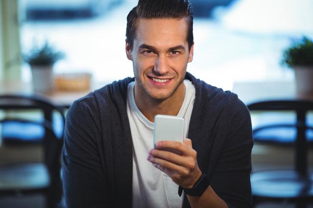 Young man sitting in a cafe, smiling while using a mobile phone. Ideal for themes related to technology, communication, modern lifestyle, and casual settings. Can be used in advertisements, blog posts, and social media content promoting mobile apps, cafes, or lifestyle products.