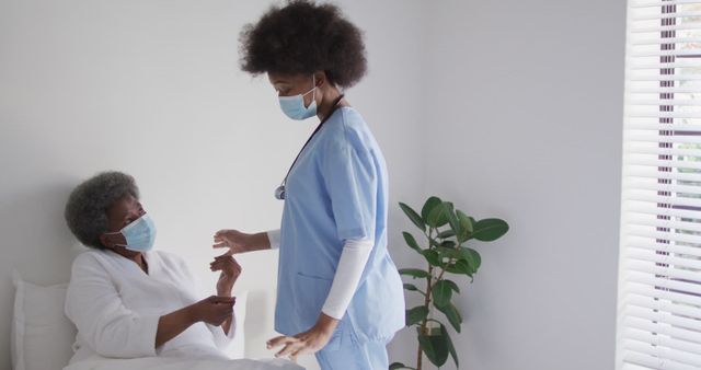 African American nurse in scrubs and mask assists senior female patient with mask in hospital room. Ideal for themes related to elderly care, healthcare professionals, COVID-19 safety measures, and medical assistance.