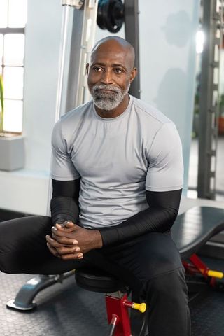 Mature african american man sitting on gym bench wearing gray shirt and arm sleeves