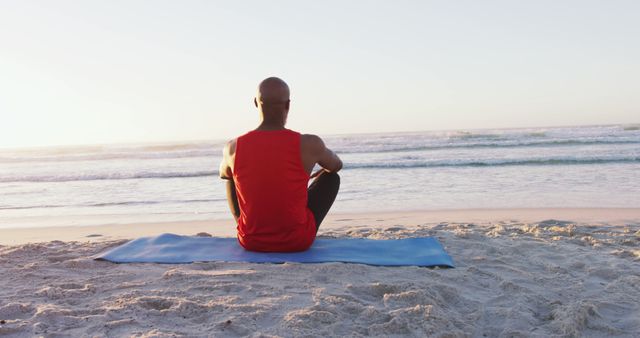 Back of man sitting on blue yoga mat on sandy beach facing the ocean. Wearing red tank top, observing sunset. Perfect for themes of relaxation, tranquility, fitness, wellness, summer retreats, and mindfulness practices.