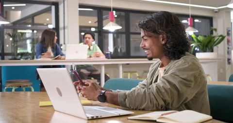 Smiling Young Professional Using Smartphone and Laptop in Modern Coworking Space