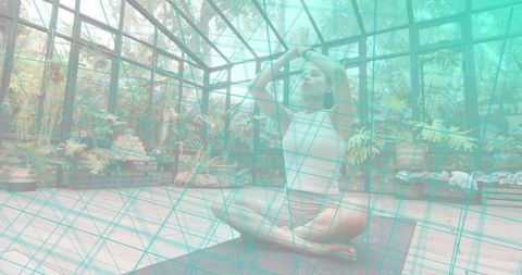 Woman practicing yoga meditation inside glasshouse surrounded by lush plants and wood