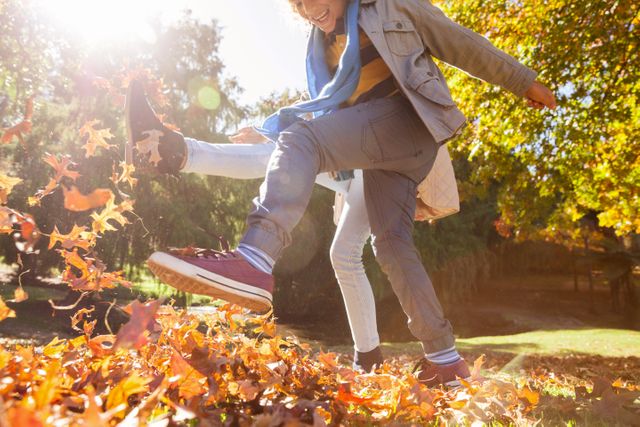 Friends enjoying a playful moment kicking autumn leaves in a park. Ideal for use in advertisements, social media posts, and articles about outdoor activities, friendship, and seasonal fun.
