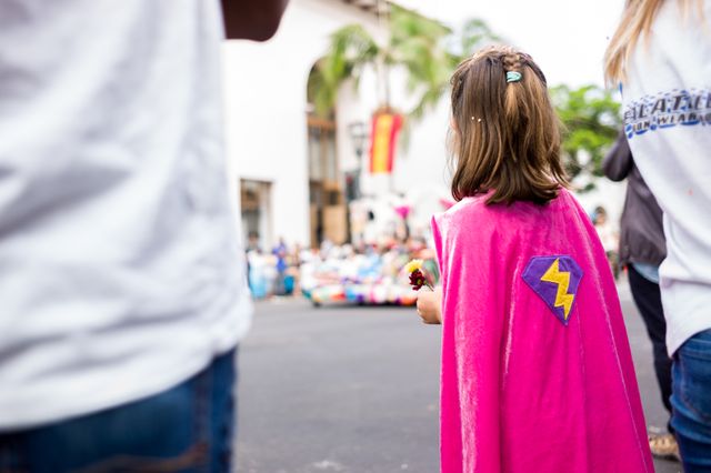 Child wearing pink superhero cape standing among crowd at a community event. Bright colors and festive atmosphere highlight themes of childhood joy and community. Ideal for use in promotions for community events, children's celebrations, or advertisements targeting families and local activities.