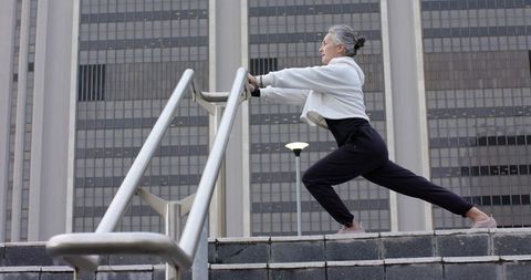 Senior woman stretching on city steps performing lunge using handrail