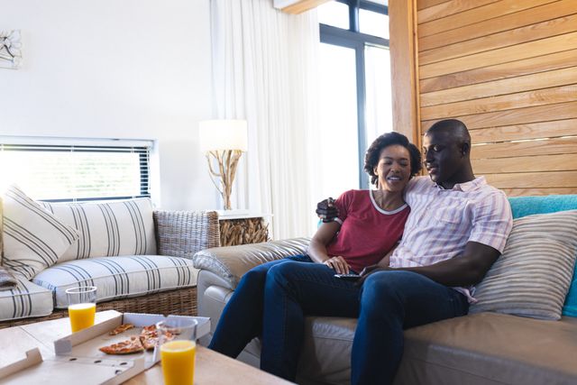 African American couple sitting on a couch in a modern living room, smiling and enjoying each other's company. Ideal for use in advertisements, blogs, or articles about relationships, home life, and family bonding. Perfect for promoting home decor, lifestyle products, or services that emphasize comfort and togetherness.