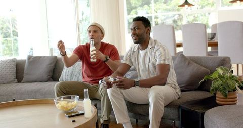 Friends playing video game and enjoying drinks and snacks on modern living room sofa