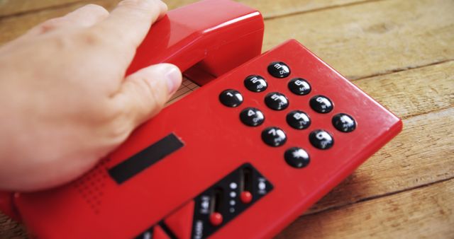 Displays a red vintage rotary telephone being picked up over a wooden desk. The image evokes nostalgia and can be used for themes related to classic retro technology, communication history, and timeless design. Suitable for blogs, articles, or advertisements focused on vintage gadgets or telecommunication topics.