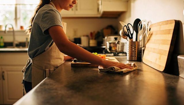 Young woman wearing beige apron wiping dark kitchen counter while preparing vegetables