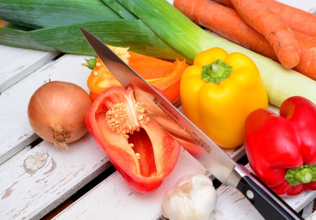 Arrangement of various fresh vegetables including peppers, onion, garlic, carrots, and a knife on wooden table. Ideal for articles on healthy eating, cooking blogs, recipe websites, or advertisements for kitchenware.