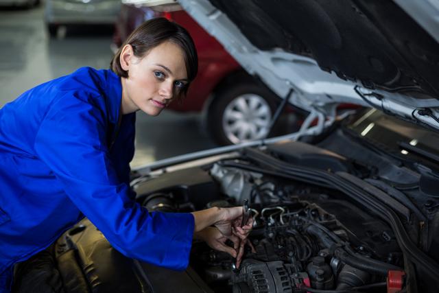 Woman in blue mechanic uniform working on car engine in automotive repair garage. Ideal for use in articles about women in automotive industry, car maintenance, professional technicians, and mechanical workshops.