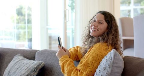 Smiling woman relaxing on gray sofa using smartphone in bright contemporary living room