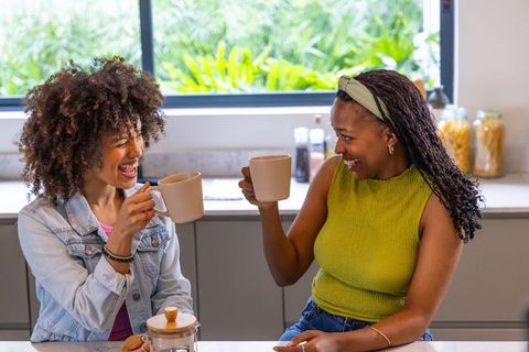 Friends toasting coffee mugs and laughing at modern kitchen counter with window view
