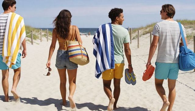 Group of friends walking along sunlit beach dunes carrying towels, flip-flops, woven tote