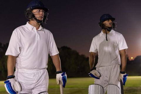 Diverse cricketers standing at dusk holding bats in white kits under stadium floodlights