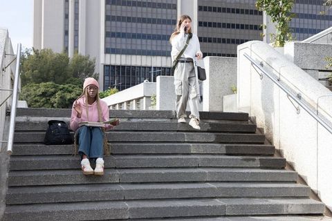 Studying and chatting on city stairs, black student sketching, white student on phone