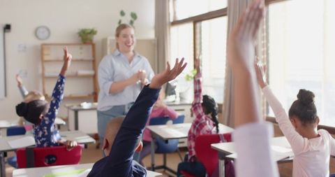 Female elementary teacher leading engaged students raising hands in happy sunlit classroom