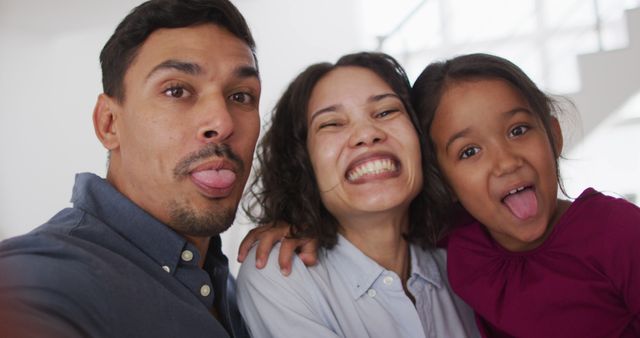 Diverse family posing for a playful selfie while sticking out their tongues. Parents and child are smiling and enjoying a moment of fun indoors. Perfect for use in family-oriented content, multicultural representation, parenting blogs, advertisements focusing on family joy, and social campaigns promoting happy family bonds.