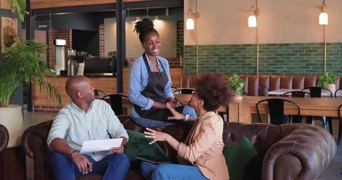 African American barista smiling and shaking hands with seated customers in cafe lounge