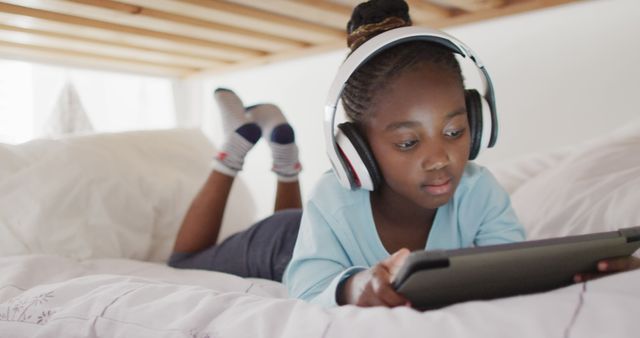 Girl lying on bed, wearing headphones, using digital tablet. Perfect for depicting technology use among children, family life, online education, entertainment, digital activities, relaxation, and casual indoor settings. Ideal for adverts, educational articles, and lifestyle blogs.