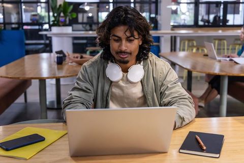 Indian man typing on laptop in modern coworking space wearing headphones, focused work