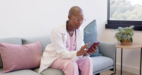 Clinician checking smartphone on sofa in pink scrubs white coat with stethoscope