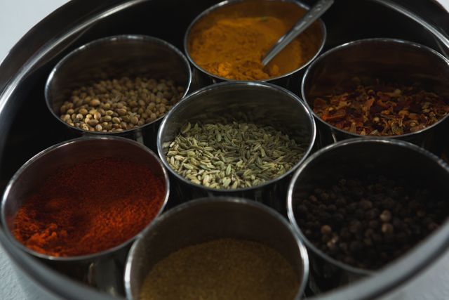 This image showcases a traditional spice box containing various spices such as turmeric, coriander, fennel, chili powder, and black pepper. Ideal for use in culinary blogs, cooking websites, recipe books, and food-related articles to illustrate the use of spices in cooking.