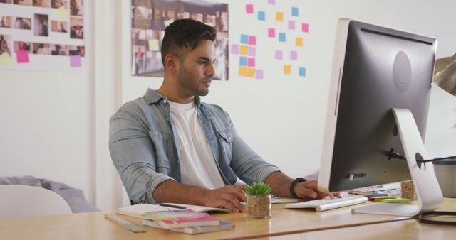 Ideal for illustrating young professionals in modern work environments, this image showcases a man at his desk using a computer. His workspace is adorned with colorful sticky notes and creative elements, suggesting a dynamic and innovative atmosphere perfect for articles on modern office setups, productivity tips, or creative process in tech startups.