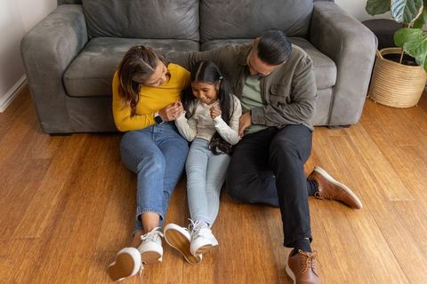 Loving family sitting on hardwood floor, parents leaning toward smiling daughter, cozy home