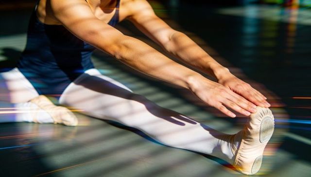 Ballet dancer stretching into seated forward split wearing leotard and tights in sunlight