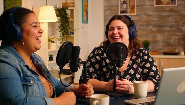 Laughing podcast hosts recording cozy conversation in home studio with microphones and headphones
