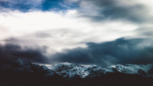 Dramatic snow-capped mountain range under stormy clouds creating a serene yet powerful landscape. Perfect for travel advertisements, nature blogs, outdoor adventure promotions, and website background images.