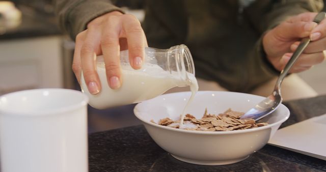 Close-up of a person pouring milk over a bowl of bran flakes in a modern kitchen. Ideal for use in advertisements or articles related to healthy eating habits, breakfast solutions, dairy products, and morning routines.