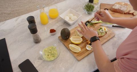 Young woman assembling avocado toast on marble countertop with eggs, lemon, fresh juice