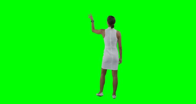 Woman in white dress points at blank green screen background. Suitable for virtual meetings, presentations, and video productions. Perfect for inclusion of custom backgrounds or digital content.