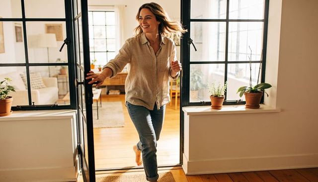Smiling woman entering sunlit home barefoot, opening black-framed door with potted plants