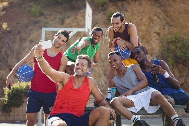 Group of basketball players taking a selfie on an outdoor court. They are smiling and appear to be enjoying their time together. This image can be used for promoting team spirit, sports events, fitness programs, recreational activities, and social bonding.