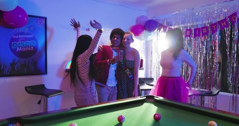 Diverse friends celebrating disco-themed 21st birthday with pool table, neon balloons