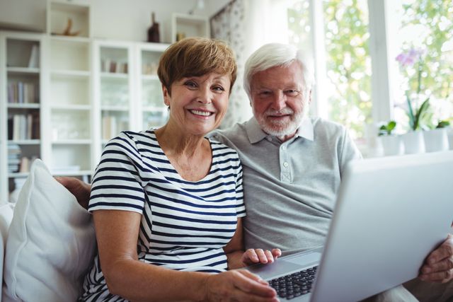 Senior couple sitting on sofa using laptop in bright living room. They are smiling and appear happy, suggesting a comfortable and relaxed environment. This image can be used for topics related to senior lifestyle, technology use among elderly, retirement, and family bonding.