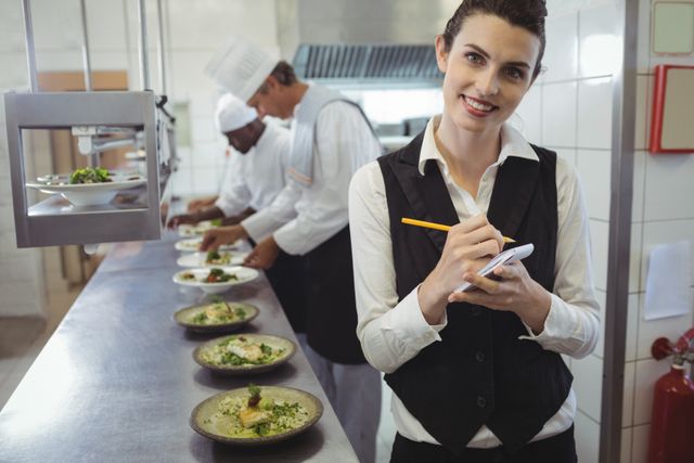Portrait of smiling waitress with notepad in commercial kitchen and chefs preparing food in background