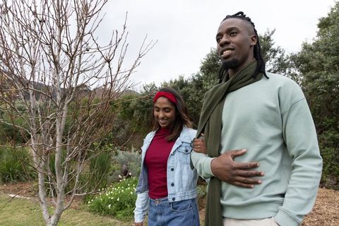 Diverse couple walking arm-in-arm through parkland wearing denim jacket and olive scarf
