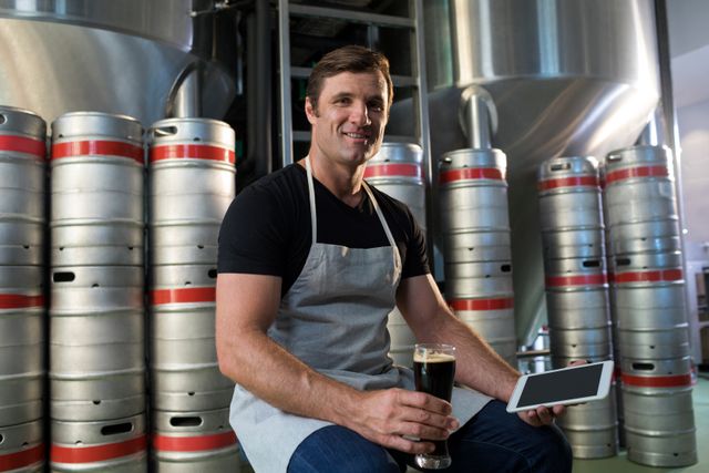 Worker in brewery holding tablet and beer while sitting by kegs. Ideal for use in articles or advertisements related to brewing, craft beer production, industrial work environments, and technology in manufacturing.